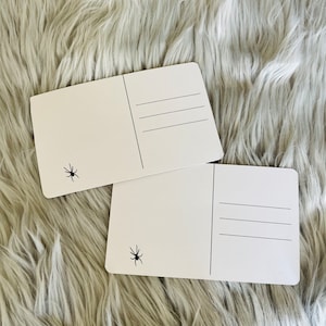 May include: Two white postcards with a black spider graphic in the corner. Each postcard has lines for the address and a vertical line for the stamp. The postcards are rectangular with rounded corners.