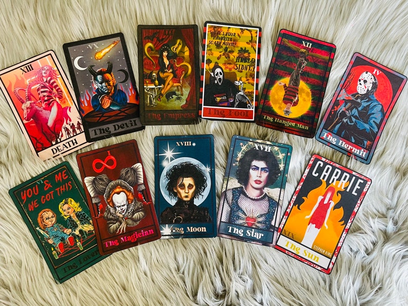May include: A collection of tarot cards featuring horror movie characters and themes. Each card has a unique design with titles like "Death," "The Devil," and "Carrie." The cards are arranged on a soft, white surface.