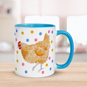 May include: A white ceramic mug with a blue rim and handle. The mug features a watercolor illustration of a brown chicken with a red comb and wattle, surrounded by colorful polka dots.