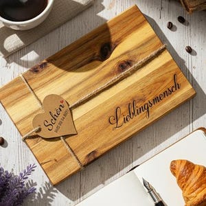 Könnte beinhalten: Ein Holzschneidebrett mit den Worten "Lieblingsmensch" und einem herzförmigen Anhänger mit der Aufschrift "Schön aus du da bist". Das Bild zeigt auch eine Tasse Kaffee, ein Croissant, ein Notizbuch und alte Bücher.