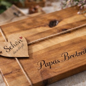 Könnte beinhalten: Ein Holzschneidebrett mit einem herzförmigen Anhänger, der mit Bindfaden gebunden ist. Der Anhänger trägt die Aufschrift "Schön dass es dich gibt" mit einem kleinen roten Herz. Das Brett ist mit "Papas Brotzeitbrett" graviert.