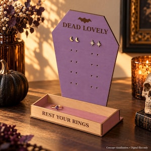 Könnte beinhalten: Ein violetter, sargförmiger Schmuckständer aus Holz mit einem Fledermaussymbol und dem Text DEAD LOVELY oben. Die Basis ist ein rechteckiges Tablett mit dem Text REST YOUR RINGS, das einen goldenen Ring mit einem violetten Stein enthält.