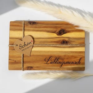 Puede incluir: Tabla de cortar de madera rectangular con una etiqueta en forma de corazón atada con hilo. La etiqueta dice "Schön, dass du da bist!" y la tabla tiene grabada la palabra "Lieblingsmensch".