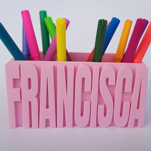 Pode incluir: Porta-lápis de plástico rosa com o nome "FRANCISCA" impresso na frente. O porta-lápis está cheio de marcadores coloridos.
