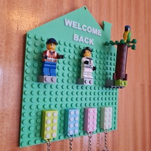 Può includere: Un portachiavi verde in stile Lego a forma di casa, con le parole "WELCOME BACK" in bianco. Presenta personaggi Lego e portachiavi colorati. Un pappagallo Lego è seduto su un albero Lego.