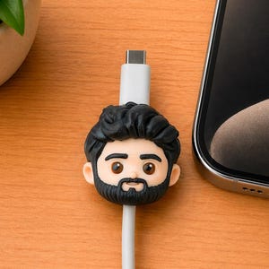 May include: A white USB-C charging cable featuring a decorative cable protector shaped like a cartoon head with black hair, a full black beard, and brown eyes. The accessory is positioned on the cable, surrounding the white plastic connector housing.