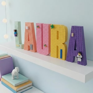 May include: The name LAURA spelled out in large, three-dimensional block letters constructed from plastic building bricks. The letters are colored light green, peach, pink, yellow, and purple, each featuring small attached figures and brick accents.