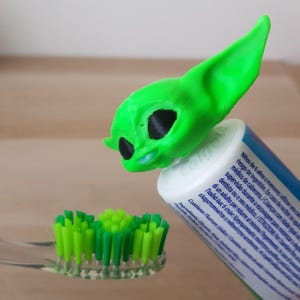 Könnte beinhalten: Eine grüne 3D-gedruckte Figur, die einem beliebten fiktiven Charakter ähnelt, ist auf der Oberseite einer weißen Zahnpastatube befestigt. Die Figur hat große Ohren und schwarze Augen. Eine grüne Zahnbürste mit grünen Borsten befindet sich im Vordergrund.