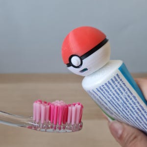 Puede incluir: Un cepillo de dientes rosa con un dispensador de pasta de dientes en forma de Poké Ball rojo y blanco.