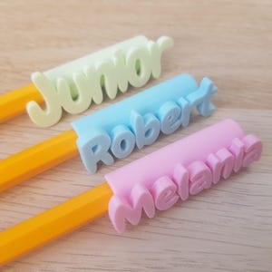 Könnte beinhalten: Drei gelbe Bleistifte mit personalisierten Namenstoppern. Die Topper sind aus Kunststoff und in den Farben hellblau, hellgrün und rosa. Die Namen auf den Toppern sind "Junior", "Robert" und "Melanie".