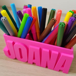 Puede incluir: Un porta lápices de plástico rosa con el nombre "JOANA" escrito en letras grandes y en relieve. El porta lápices está lleno de rotuladores de colores.