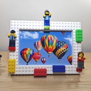 Pode incluir: Moldura branca estilo Lego com uma impressão de balões de ar quente coloridos. A moldura é decorada com blocos Lego de várias cores e minifiguras Lego. A imagem mostra um céu azul com vários balões de ar quente.