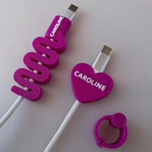 Pode incluir: Dois cabos de carregamento USB-C brancos com protetores de cabo magenta impressos em 3D. Um protetor tem formato de ziguezague e o outro de coração, ambos com o nome CAROLINE em branco. Um clipe de cabo circular magenta está incluído.