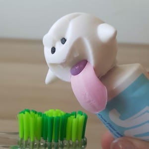 Pode incluir: Um dispensador de pasta de dentes branco em forma de fantasma com uma língua rosa que se projeta. O dispensador está preso a um tubo de pasta de dentes azul e branco. Uma escova de dentes verde está em primeiro plano.
