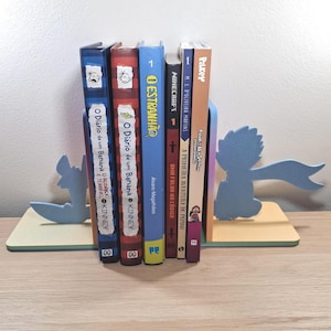 Pode incluir: Um conjunto de suportes de livros de madeira azuis e amarelos com um design lúdico, segurando uma coleção de livros coloridos. Os suportes de livros apresentam uma silhueta de um personagem com um cachecol. Os livros têm títulos em português e inglês.
