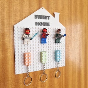 Könnte beinhalten: Ein weißes, hausförmiges Wanddisplay mit dem Text SWEET HOME in grauen Buchstaben. Das Board verfügt über ein Raster aus Noppen mit drei befestigten Superhelden-Minifiguren und drei bunten Schlüsselanhängern aus Bausteinen an Metallringen.