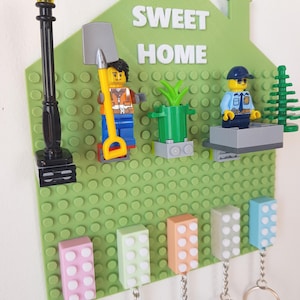 Può includere: Un supporto da parete a forma di casa verde chiaro con la scritta bianca SWEET HOME. Include un lampione nero, una minifigure di operaio con una pala, una pianta verde, una minifigure di poliziotto e cinque ganci portachiavi colorati.