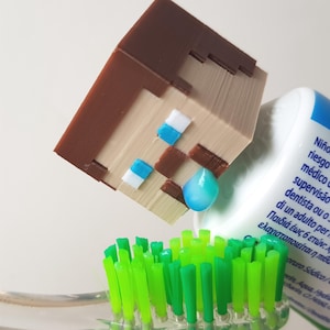 Può includere: Una figurina di Minecraft stampata in 3D di colore marrone e beige, che spreme dentifricio blu su uno spazzolino da denti verde.