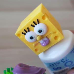 Pode incluir: Um personagem amarelo de esponja com uma expressão engraçada, incluindo uma língua roxa que se projeta, e grandes olhos brancos com pupilas pretas. O personagem é um brinquedo impresso em 3D.