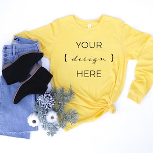 May include: A yellow long-sleeved t-shirt with the text "YOUR {design} HERE" on the front. The shirt is laid flat on a white background with a pair of blue jeans and black ankle boots.