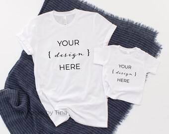 May include: Two white t-shirts with the text "YOUR {design} HERE" on them. The shirts are laid out on a blue and white striped fabric.