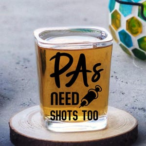 May include: A clear glass shot glass filled with an amber liquid. The front of the glass features black text that reads PAs NEED SHOTS TOO, with a small graphic of a medical syringe replacing the letter O in the word SHOTS.