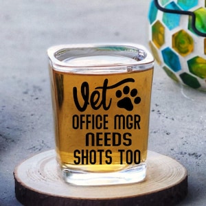May include: A square shot glass filled with a golden liquid, resting on a wooden coaster. The glass has black text that reads "Vet OFFICE MGR NEEDS SHOTS TOO" with a paw print. A blue towel and a decorative lantern are in the background.