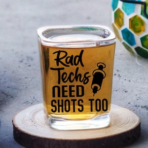 May include: A square shot glass filled with a golden liquid, resting on a wooden coaster. The glass has black text that reads "Rad Techs NEED SHOTS TOO" with a syringe graphic. A blue towel with white anchor designs is visible.