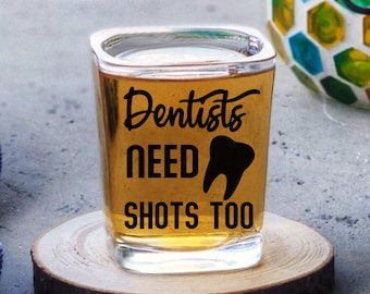 May include: A square shot glass filled with a golden liquid, resting on a wooden coaster. The glass has the text "Dentists NEED SHOTS TOO" with a tooth graphic. A blue towel with white anchor designs is on the left.