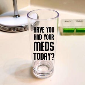 May include: A clear shot glass with the text "HAVE YOU HAD YOUR MEDS TODAY?" in black. A pill organizer with green compartments is visible in the background. A small, round mirror is also in the background.