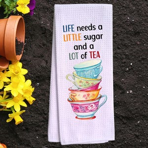 May include: White kitchen towel with the text "LIFE needs a LITTLE sugar and a LOT of TEA" and a stack of colorful teacups. Yellow daffodils and a terracotta pot are in the background.