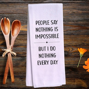 May include: A white kitchen towel with the text "People Say Nothing Is Impossible But I Do Nothing Every Day." Two wooden salad spoons are tied together with a raffia bow. Yellow and orange flowers are scattered around the towel.