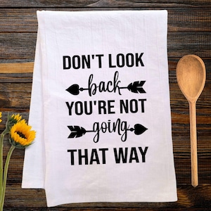 May include: White kitchen towel with the text "DON'T LOOK back YOU'RE NOT going THAT WAY" in black. A wooden spoon and sunflowers are next to the towel on a wooden surface.