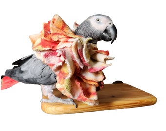 Puede incluir: Una escultura realista de un loro gris con cola roja, posado sobre una base de madera. El loro lleva un collar colorido y con volantes en tonos rojos, naranjas y amarillos, que se asemeja a una flor.