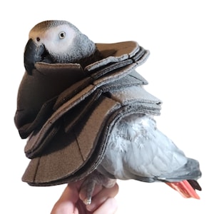 May include: A gray parrot wearing a brown fleece bird harness with multiple layers. The harness is designed to help keep the bird safe and secure.