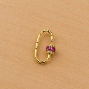 May include: A gold-colored oval-shaped clasp with a cylindrical center adorned with rectangular, deep red gemstones. The clasp's center is also embellished with small, clear gemstones. The background is a light brown wooden surface.