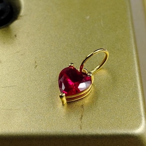 May include: A heart-shaped pendant with a vibrant red gemstone. The pendant is set in a gold-coloured metal and features a loop for a chain. The gemstone is faceted, catching the light. Jewellery.