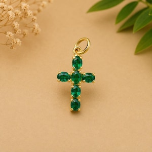 Puede incluir: Colgante de cruz dorada adornado con piedras preciosas verdes esmeralda de corte ovalado. La cruz tiene un lazo en la parte superior para una cadena. Las piedras están dispuestas simétricamente, creando un diseño clásico.