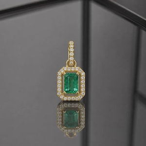 Può includere: Un pendente in oro con una gemma verde taglio smeraldo, circondata da un alone di piccole gemme trasparenti. Il pendente ha un'ansa abbinata ed è esposto su una superficie riflettente.