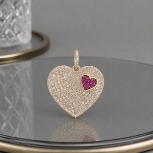 May include: A gold heart-shaped pendant featuring a pavé setting of numerous small clear stones covering the surface, with a smaller, distinct heart-shaped accent inlaid on the right side, encrusted with vibrant pink stones and a gold bail.