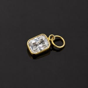 May include: A rectangular pendant featuring a clear, faceted gemstone set in a polished gold-toned bezel. The pendant includes a small, circular gold-toned jump ring attached to one side, designed for easy connection to a chain or bracelet.