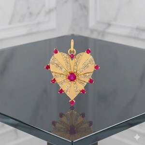 May include: A gold heart-shaped pendant featuring a central round red gemstone, surrounded by radiating rays of small clear pavé-set stones. The edges of the heart are accented with additional round red gemstones at each point and a small bail.