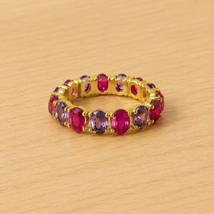 May include: A gold-toned ring featuring alternating oval-cut gemstones in shades of magenta and purple. The ring is set on a light-colored wooden surface, showcasing the vibrant colors of the stones.