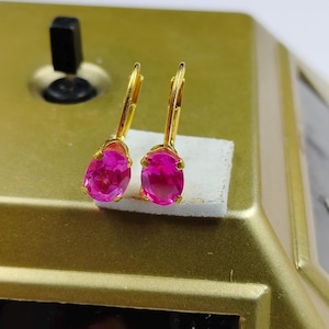 Puede incluir: Un par de pendientes dorados con cierre de palanca, cada uno con una vibrante gema rosa intenso de forma ovalada. Los pendientes se exhiben sobre una superficie dorada, mostrando su color y diseño.