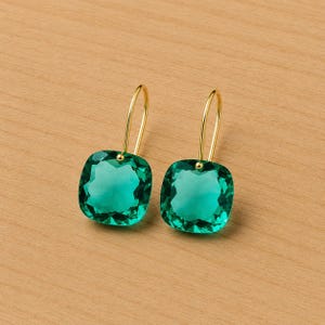 Könnte beinhalten: Ein Paar goldfarbene Ohrhänger mit großen, quadratisch geschliffenen, smaragdgrünen Edelsteinen. Die Ohrringe haben ein schlichtes, elegantes Design mit einem goldenen Haken, der sich über das Ohr wölbt.