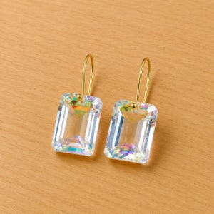 May include: A pair of gold-coloured earrings featuring rectangular, clear, faceted crystals. The crystals have a rainbow iridescence. The earrings are displayed on a light brown wooden surface.