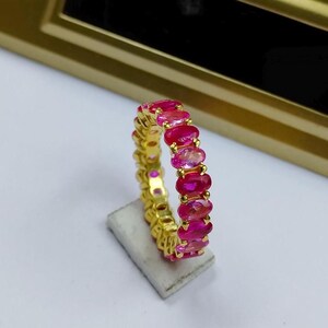 May include: A gold-colored ring featuring a continuous row of oval-cut, vibrant pink gemstones. The ring is set on a small, rectangular, light-colored stand, with a gold-colored background.