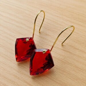 May include: A pair of red, geometric earrings with gold-colored hooks. Each earring features a faceted, diamond-shaped red gem with a small gold accent. The earrings are displayed on a light-colored wooden surface.