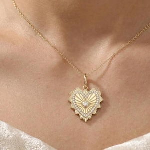 May include: A gold-colored necklace with a heart-shaped pendant. The pendant features a sunburst design with a central diamond and is surrounded by smaller diamonds. The necklace is displayed on a person's neck.