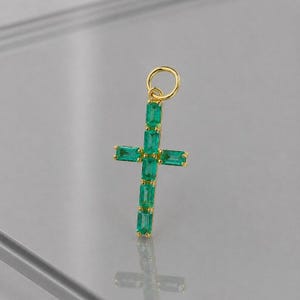 May include: A gold-coloured cross pendant, adorned with emerald-green rectangular gemstones. The cross features a loop for a chain. The gemstones are arranged in a vertical and horizontal pattern, forming a religious symbol.